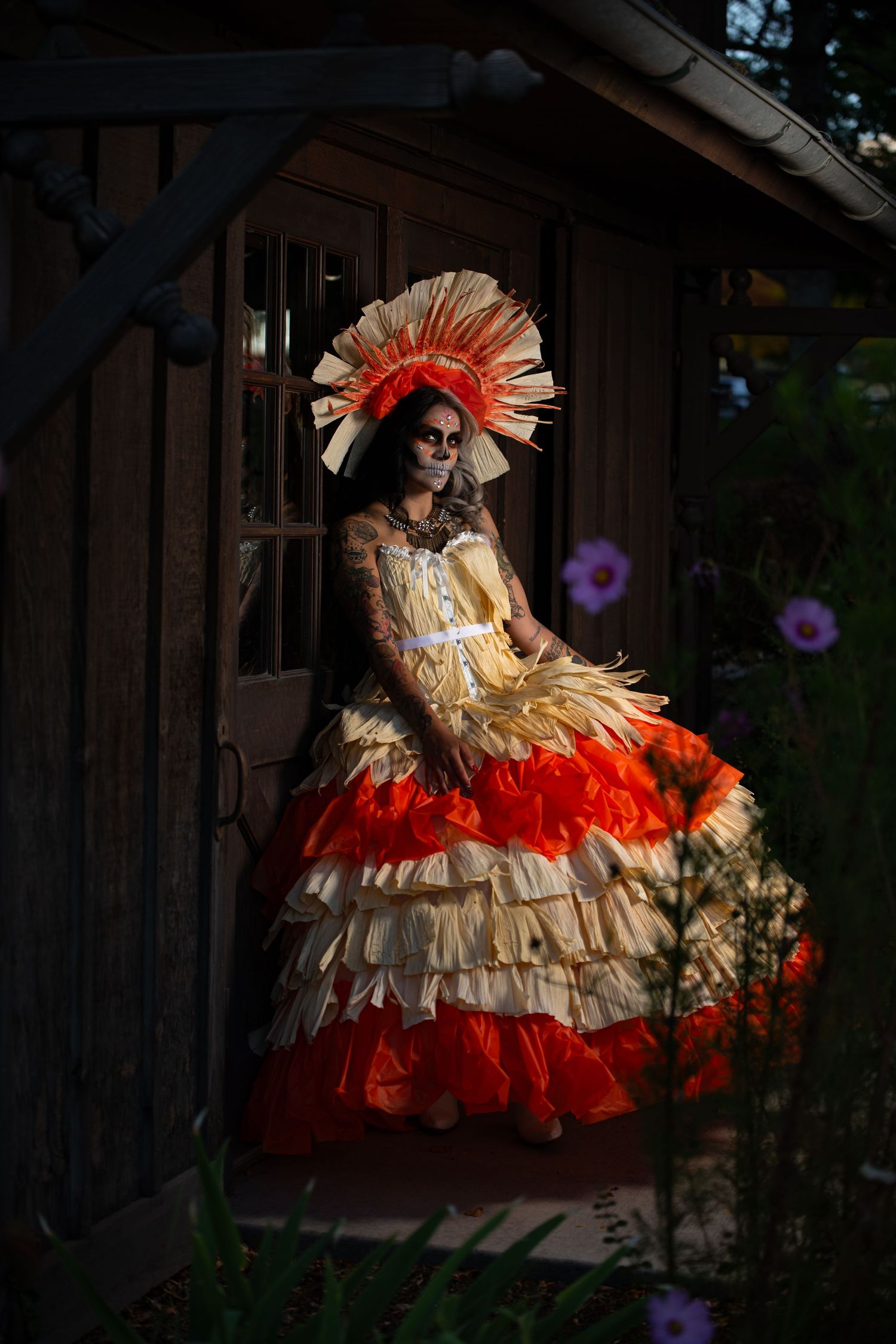 Woman in Day of the Dead costume with skull makeup and orange ruffled dress posing by rustic wooden door.
