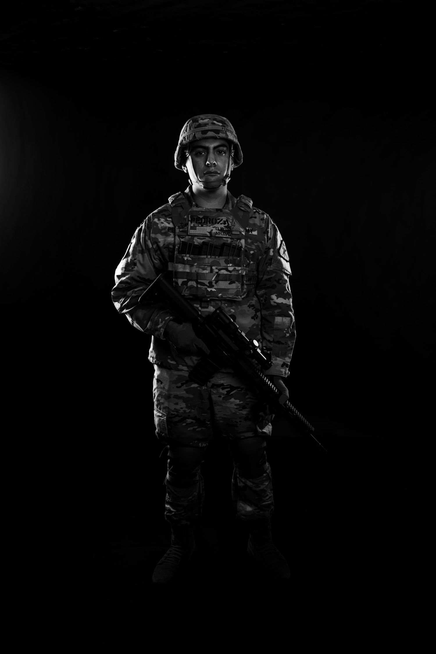 Black and white full-length portrait of US Army soldier in camouflage uniform holding rifle on dark background.