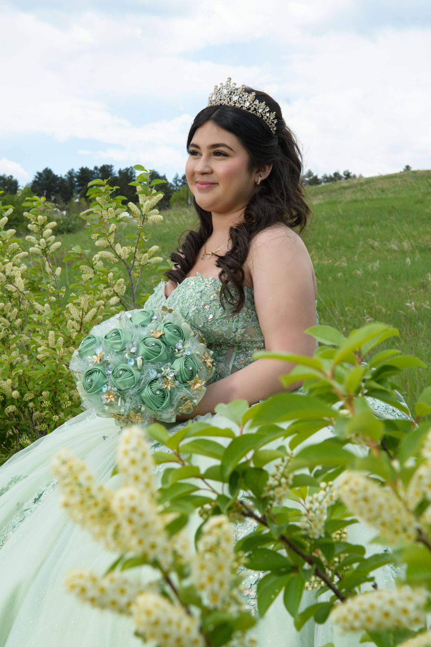 Young woman in mint green quinceañera ball gown with tiara holding bouquet in meadow with flowers.