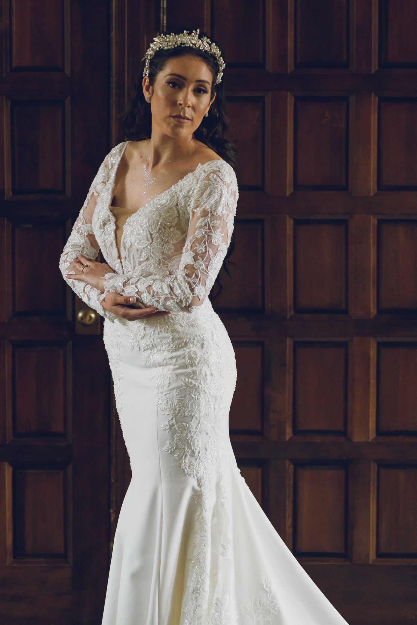 Elegant bride in fitted long-sleeve lace wedding gown with floral crown poses before wooden doors.