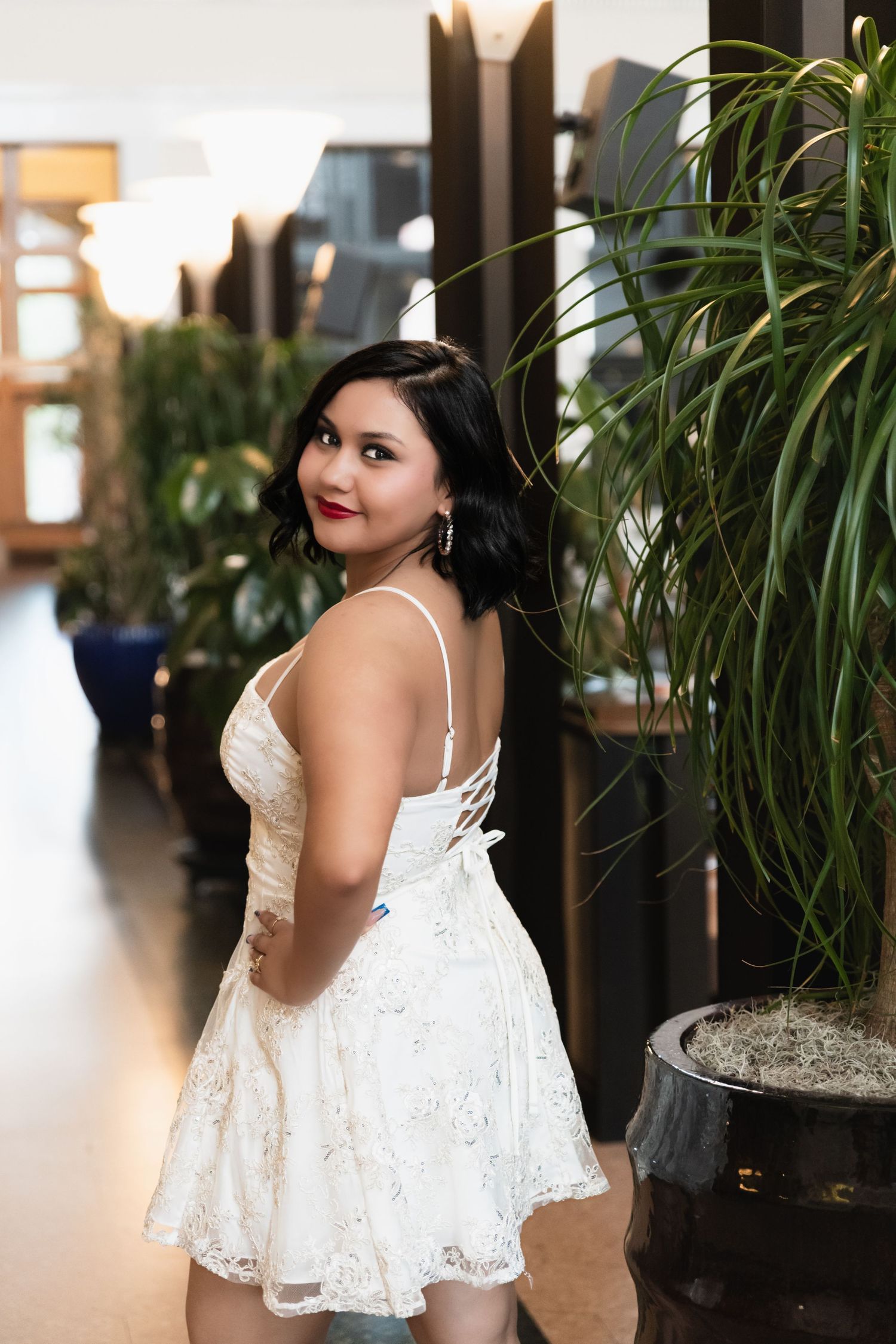 Young woman in white lace dress posing indoors near tropical plants, looking over her shoulder.