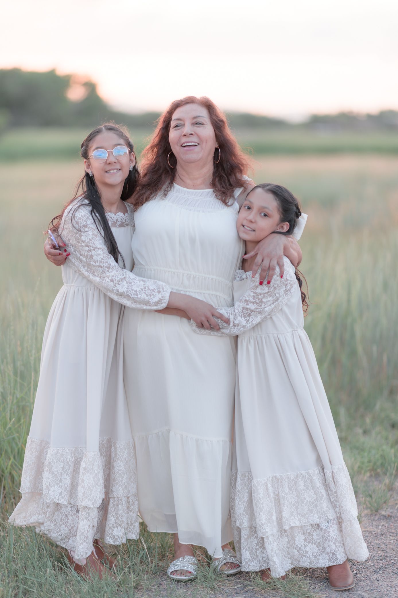 A woman with red hair and two girls in matching white dresses embrace outdoors in a field at sunset.