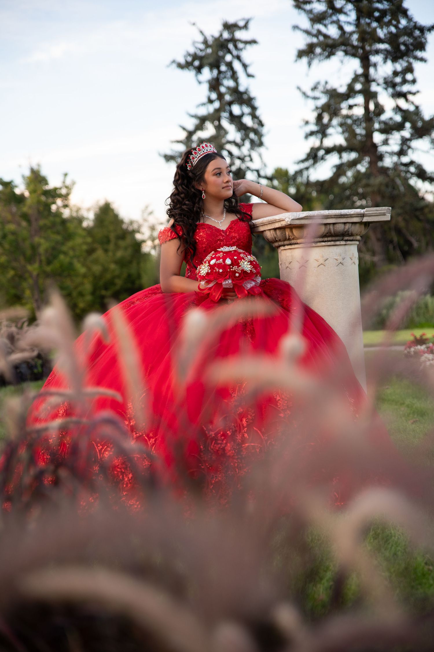 Elegant young woman in red quinceañera gown with tiara posing by stone pillar in garden setting.
