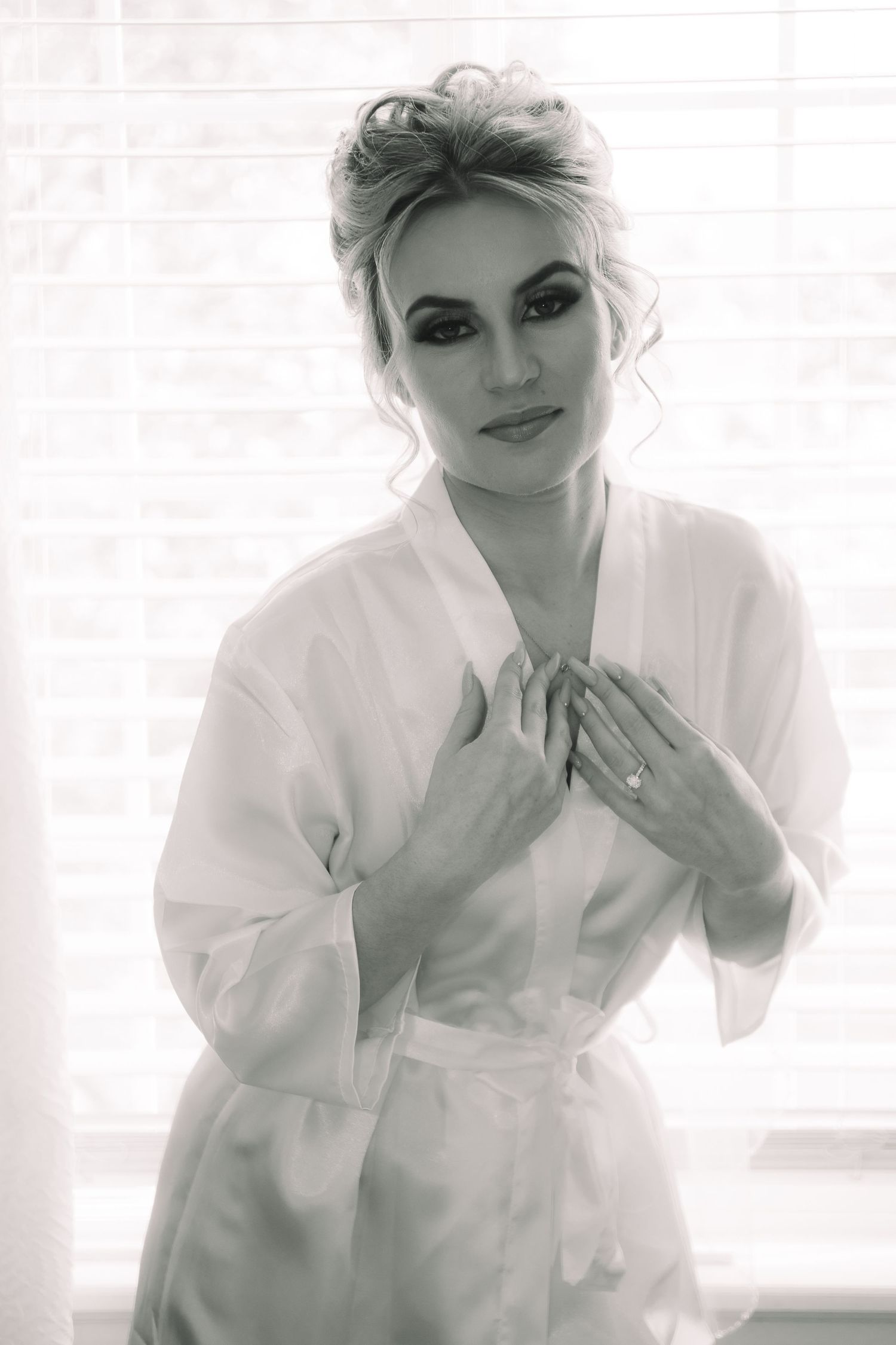 Black and white portrait of bride in white robe with dramatic makeup holding robe closed at window.