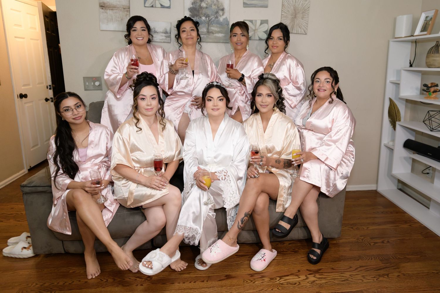 A group of bridesmaids wearing pink and white satin robes posing together indoors holding drinks before a wedding.