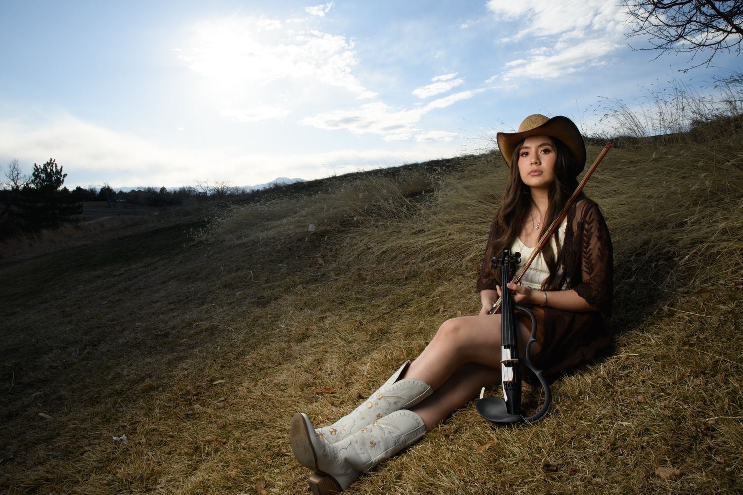 Young woman in cowboy hat and white boots sitting on a grassy hillside holding a guitar outdoors.