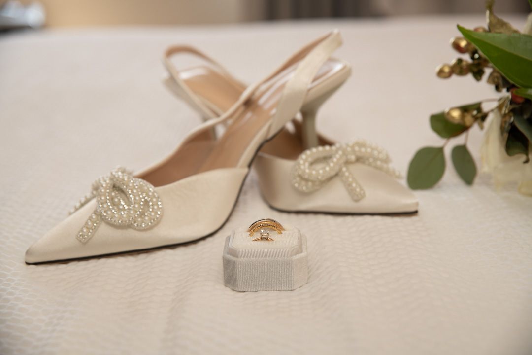 White satin bridal heels with pearl bow detail beside gold wedding rings on white velvet ring box.