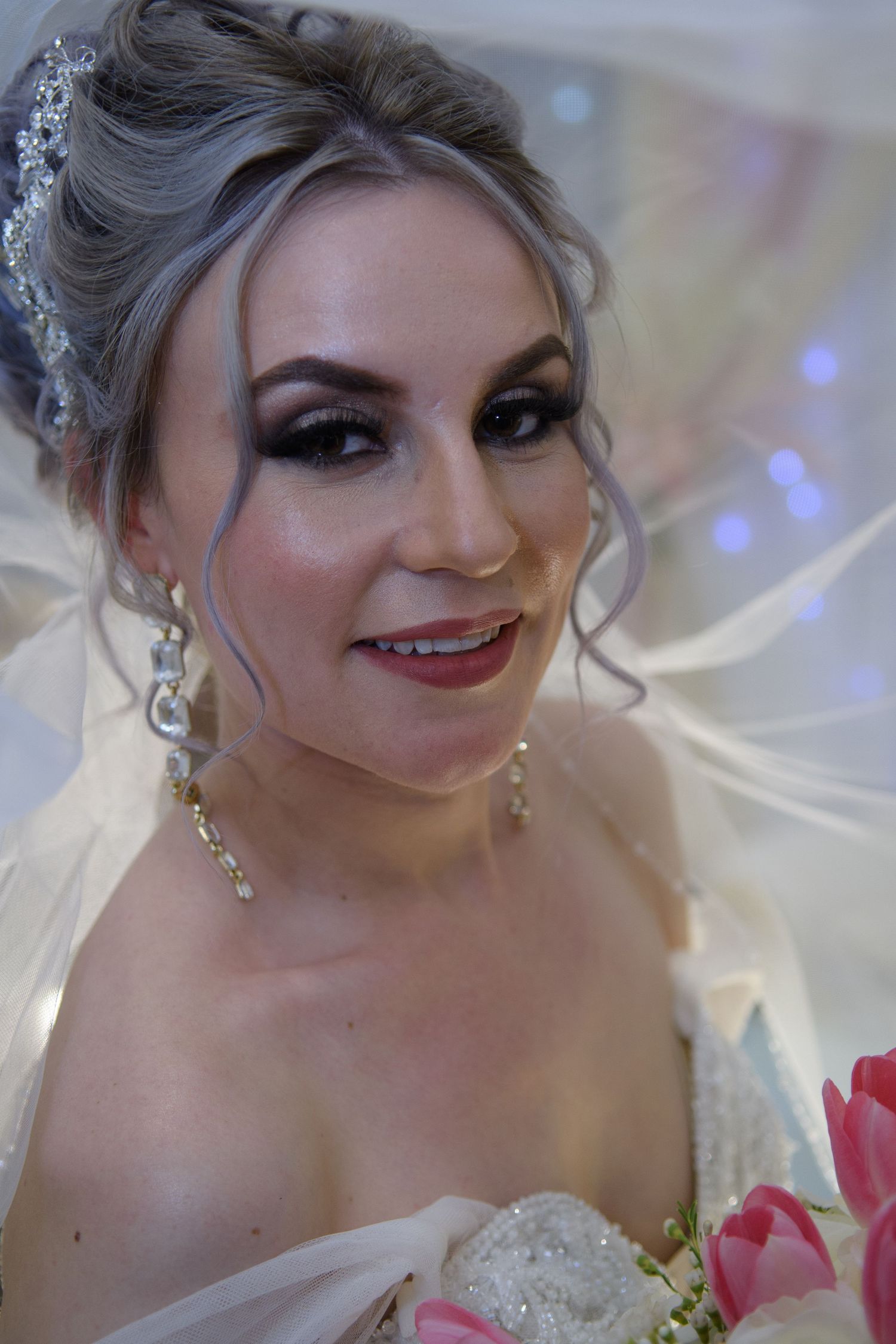 Close-up portrait of bride with silver updo, dramatic eye makeup, crystal earrings, and pink bouquet.