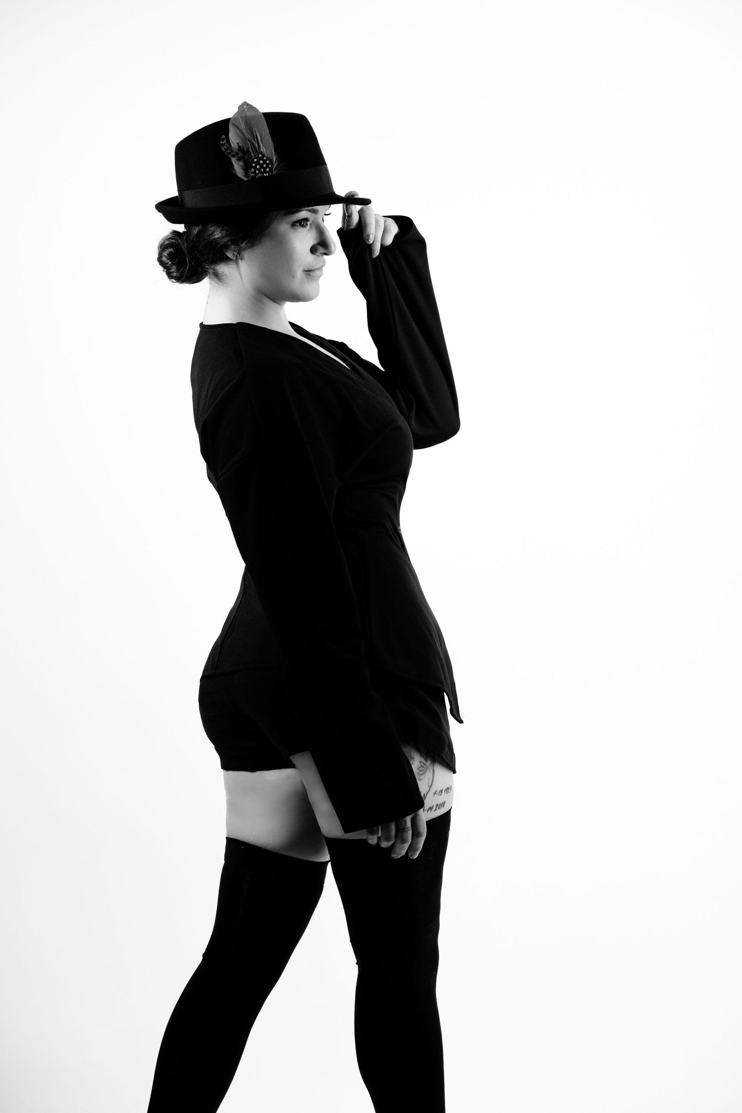 Elegant woman in black blazer, hat, and thigh-high stockings poses in dramatic black and white studio portrait.