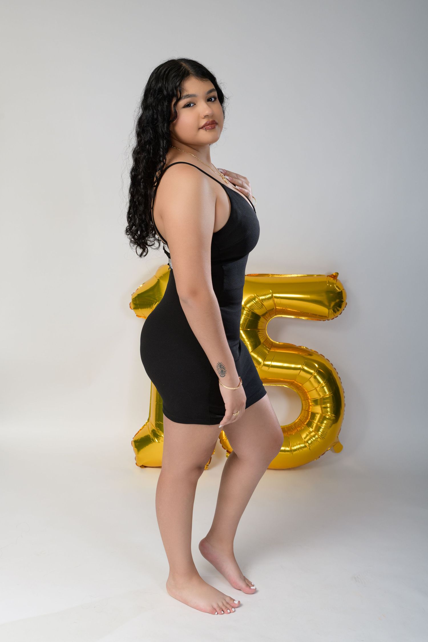 Young woman in black mini dress posing beside gold number 15 balloon on white background.