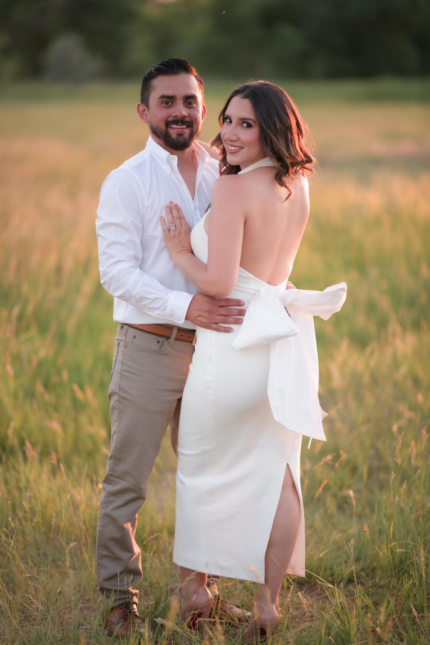 Happy couple embracing in golden field at sunset, woman in backless white dress.