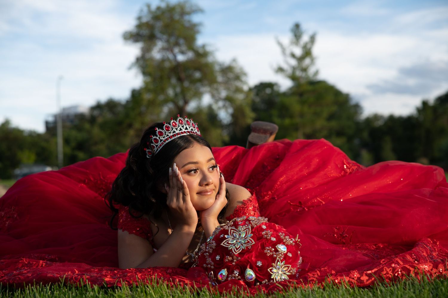 Young woman in red quinceañera ball gown with tiara lying on grass holding floral bouquet outdoors.