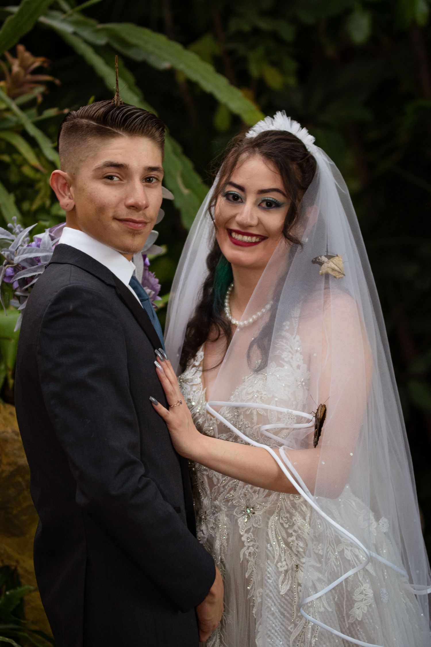 Bride and groom posing together outdoors, bride wearing lace wedding dress with veil and pearl necklace.