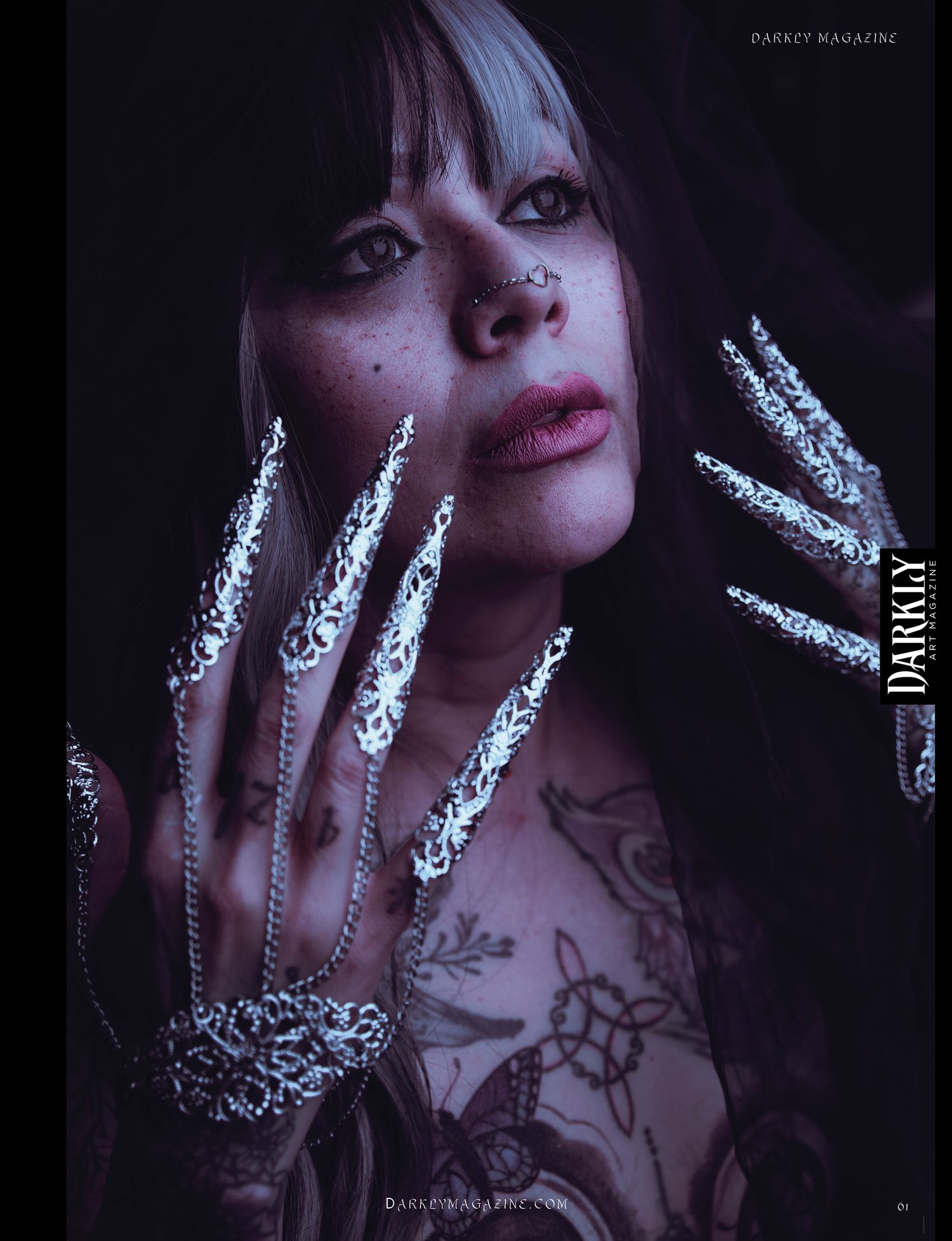 Gothic portrait of a tattooed woman with a nose ring and dark hair, wearing long ornate silver claw nail rings and a hand harness bracelet.