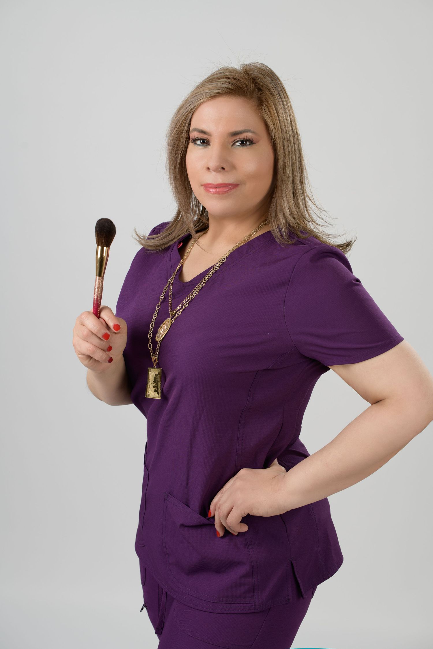 Woman in purple scrubs holding a makeup brush, wearing gold necklaces, posing confidently against a gray background.