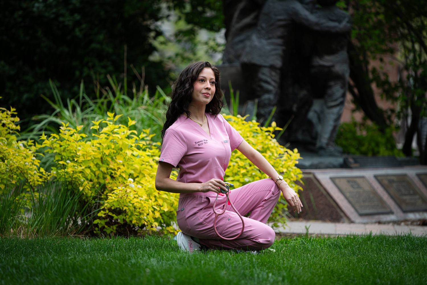 Woman in pink scrubs kneeling on grass near yellow flowers and bronze statue in outdoor garden.