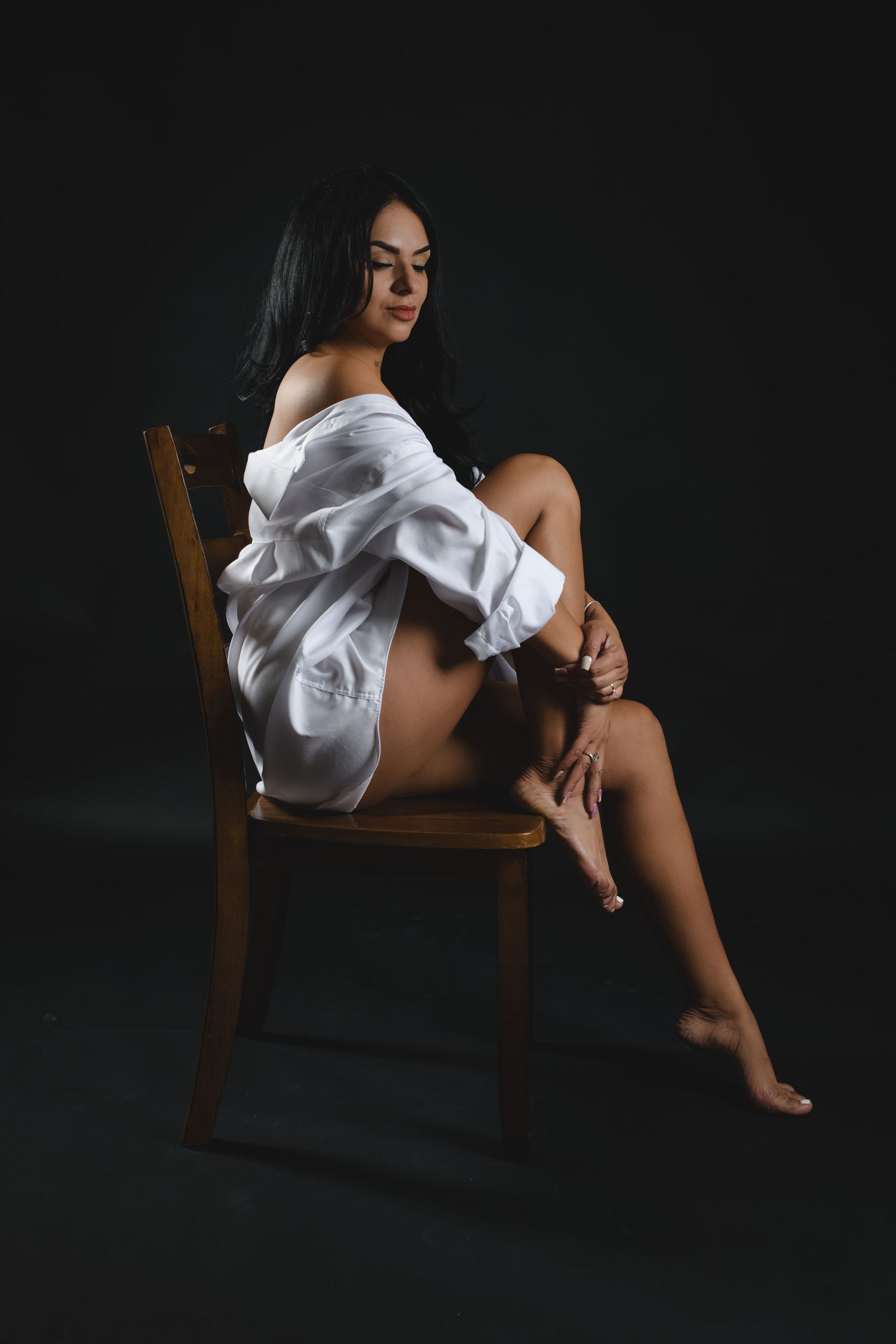 Young woman in white off-shoulder shirt seated on wooden chair against dark background.