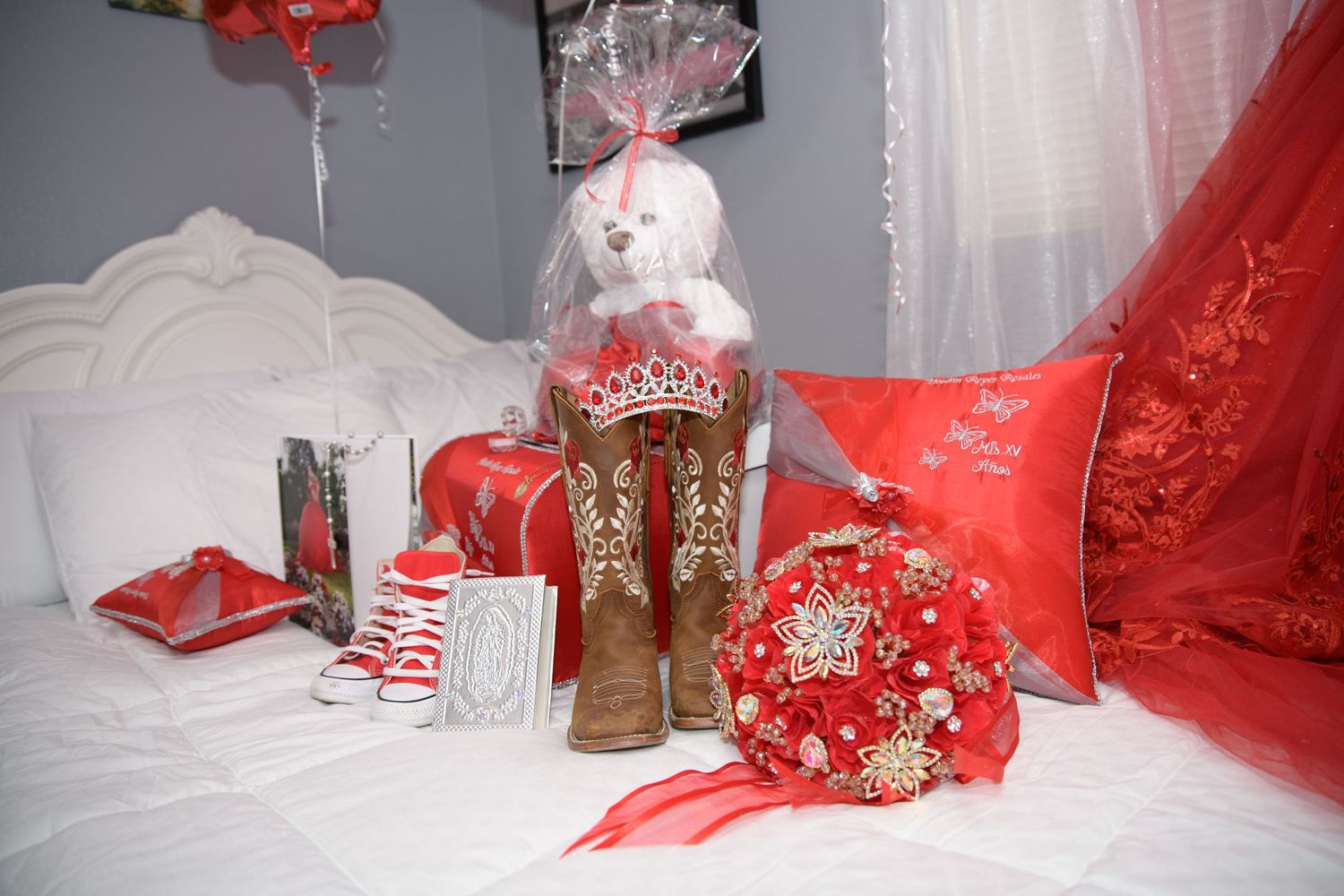 Red quinceañera themed decorations arranged on a bed including cowboy boots, bouquet, tiara bear, and pillows.