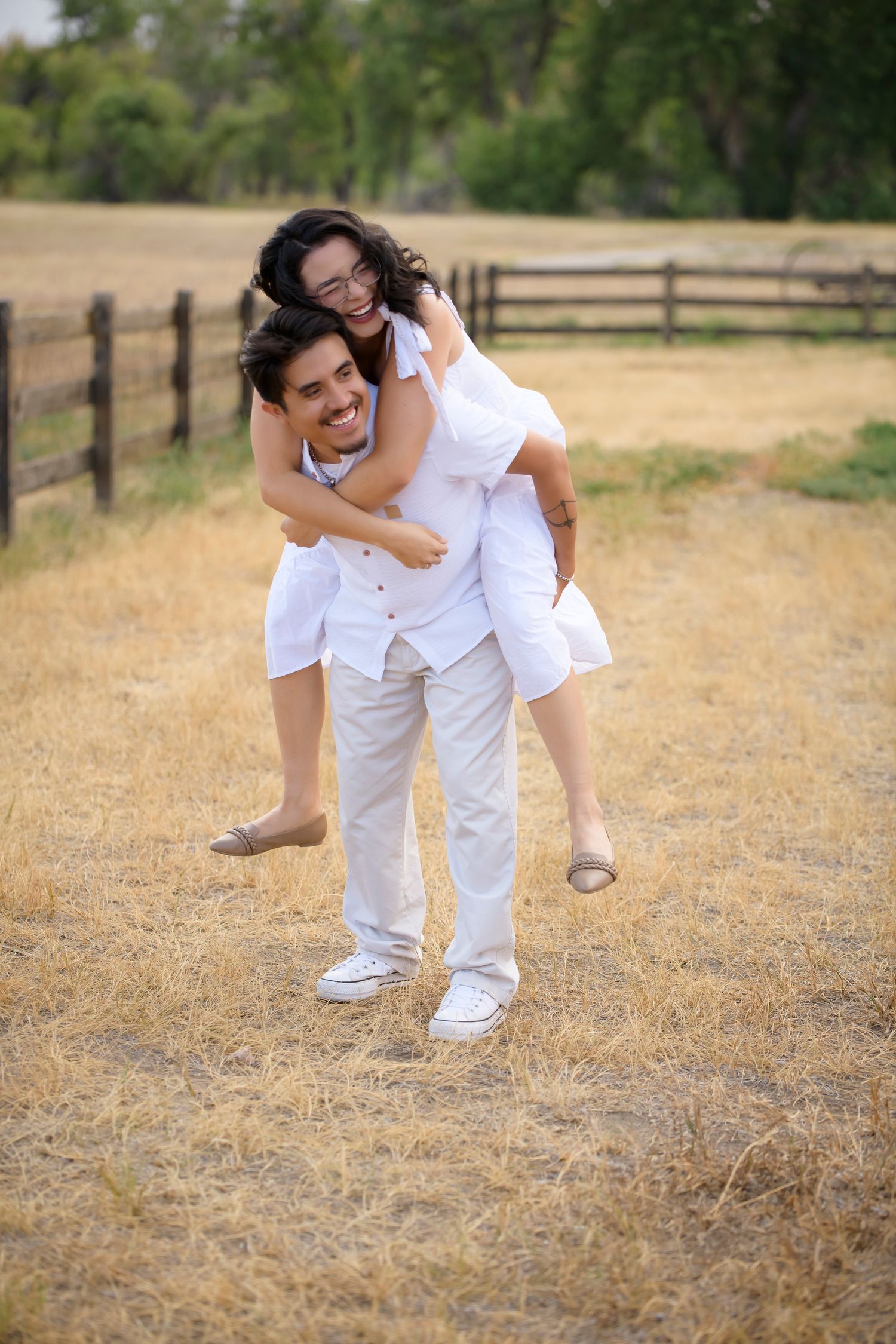 A joyful couple in matching white outfits shares a piggyback ride in a sunny field with a wooden fence backdrop.