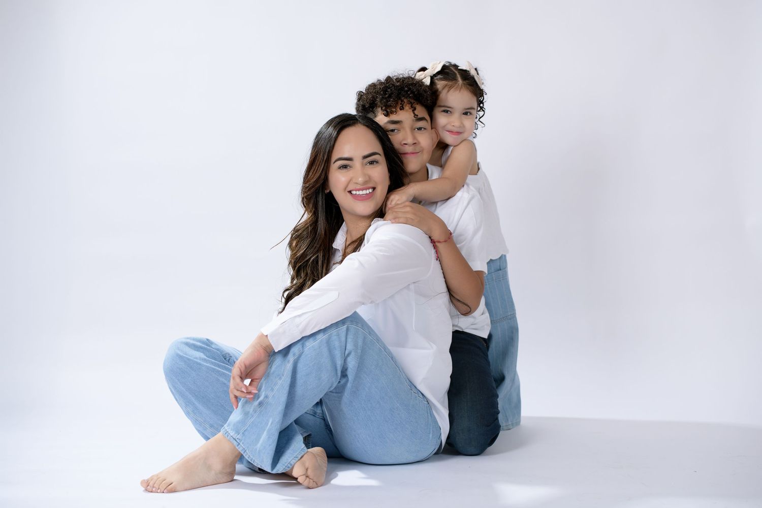 A woman and two children pose together smiling in matching white tops and blue jeans against a white background.
