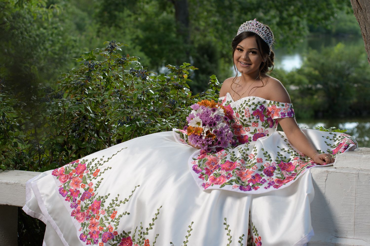Young woman in white floral quinceañera gown with tiara holds colorful bouquet while seated outdoors.