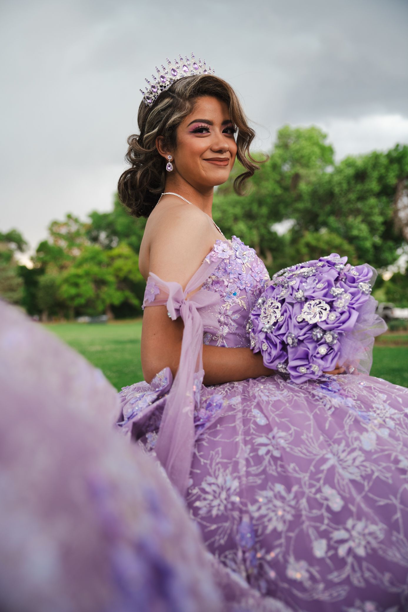 Smiling quinceañera in lavender off-shoulder gown with crystal tiara holding purple bouquet.