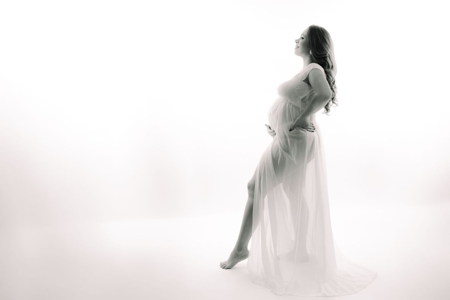 Black and white maternity photo of a pregnant woman in flowing white gown in a bright studio.