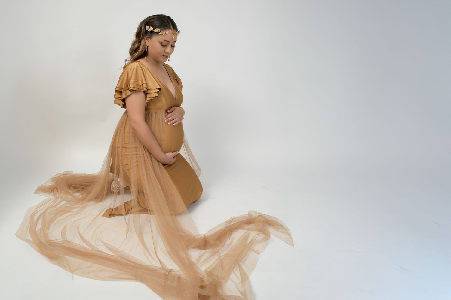 Pregnant woman in golden tulle maternity gown with floral crown posing gracefully in white studio.