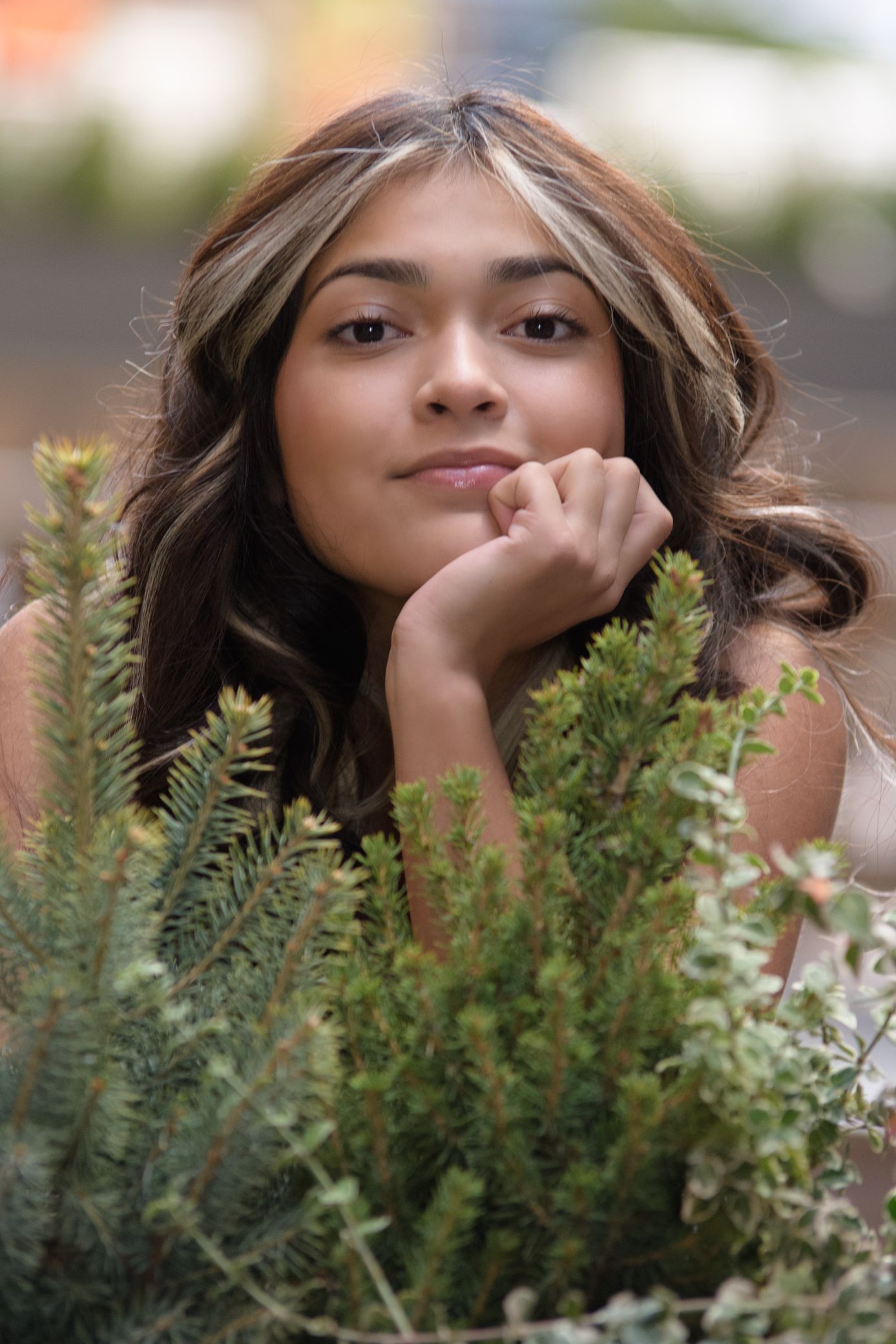 Young woman with highlighted hair resting chin on hand among green pine plants, smiling softly.