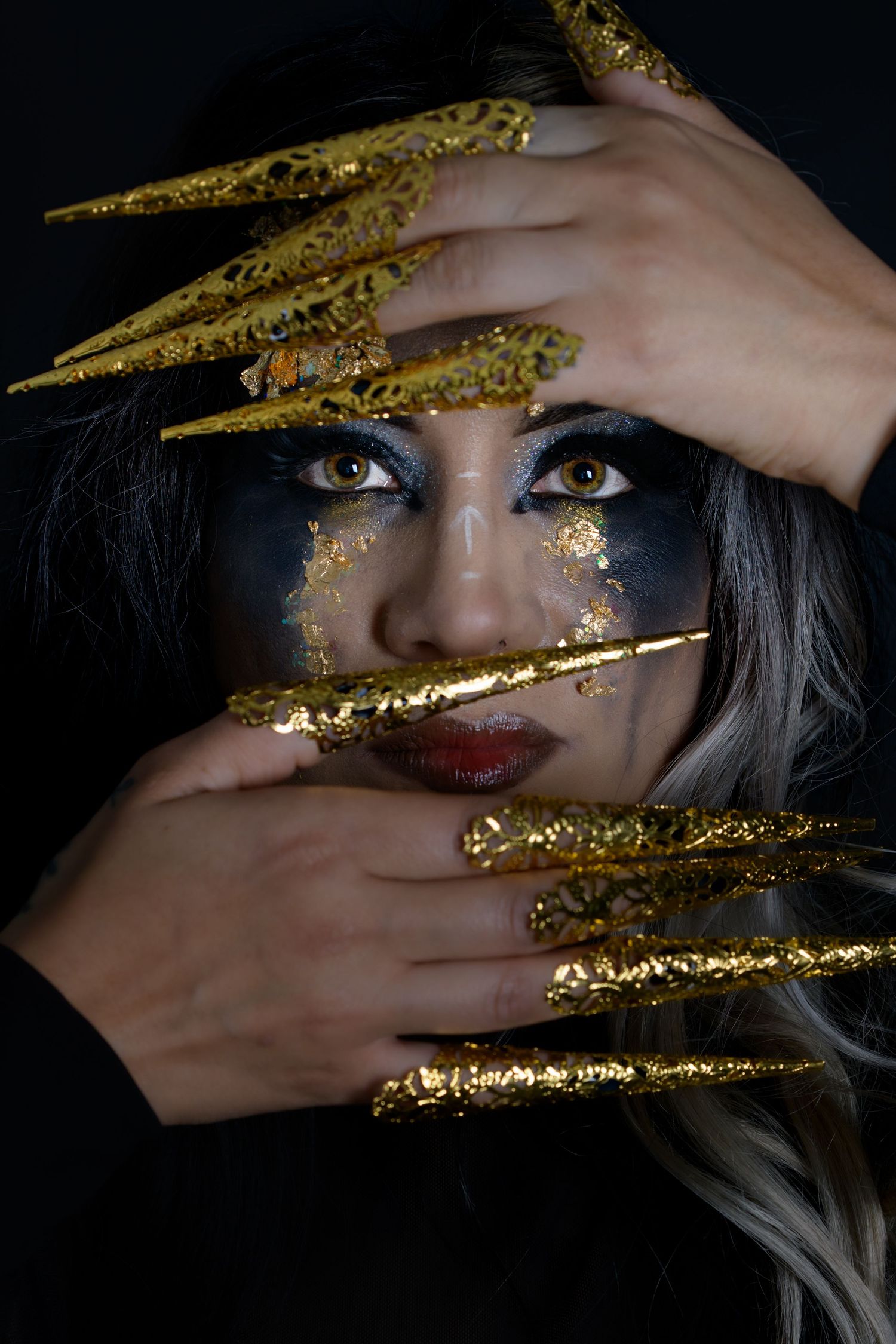 Woman with dramatic gold face paint and long gold claw nails poses against a dark background.