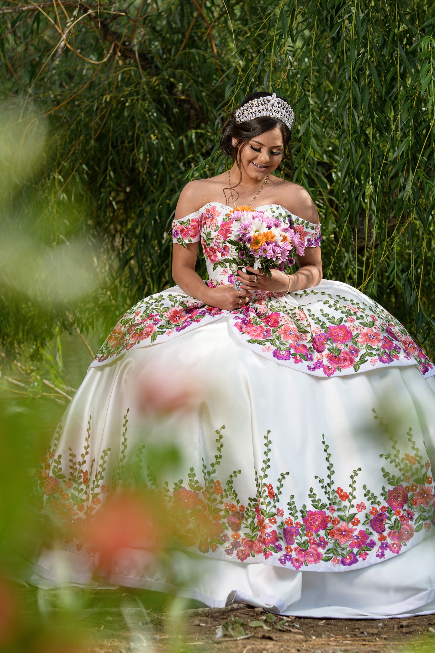 Young woman in white floral embroidered quinceañera gown with tiara holding colorful bouquet.