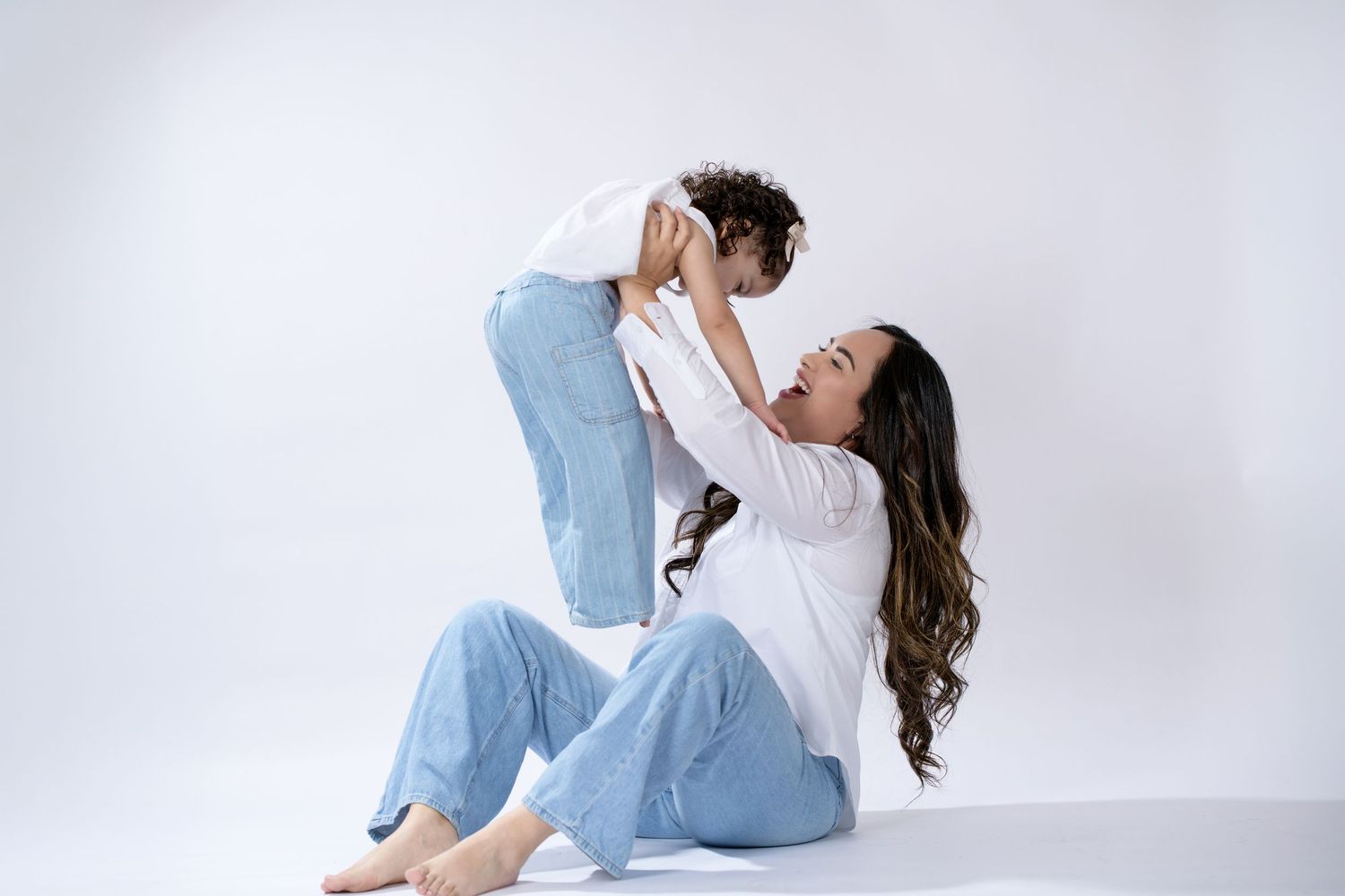 A mother sits on the floor lifting her toddler joyfully, both wearing white tops and blue jeans on a white background.