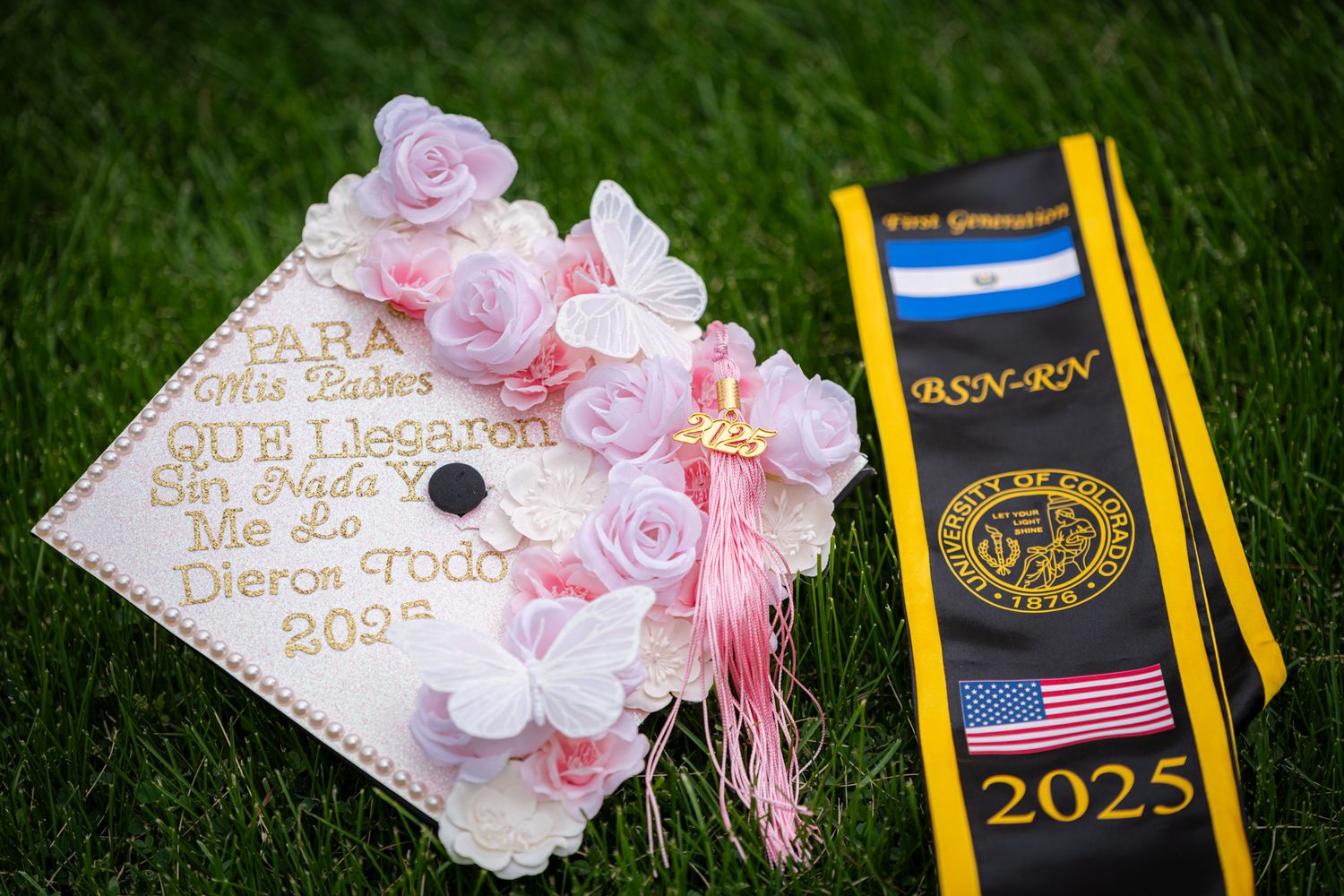 Decorated pink graduation cap with flowers and Spanish dedication message beside BSN-RN stole on green grass.