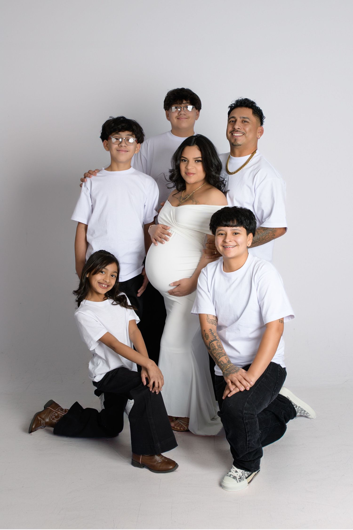 Pregnant woman in white gown poses with family of four children and partner in matching white tees for maternity photo.
