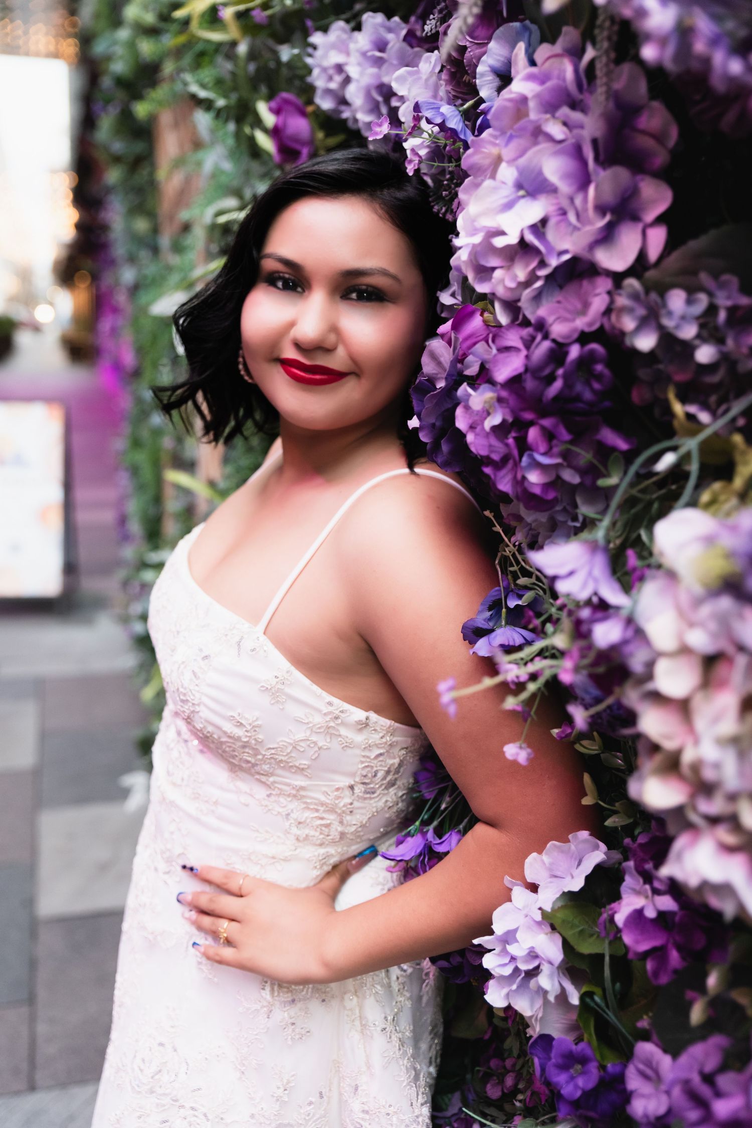 Woman in white lace dress posing beside purple floral arch, smiling with red lips and dark hair.