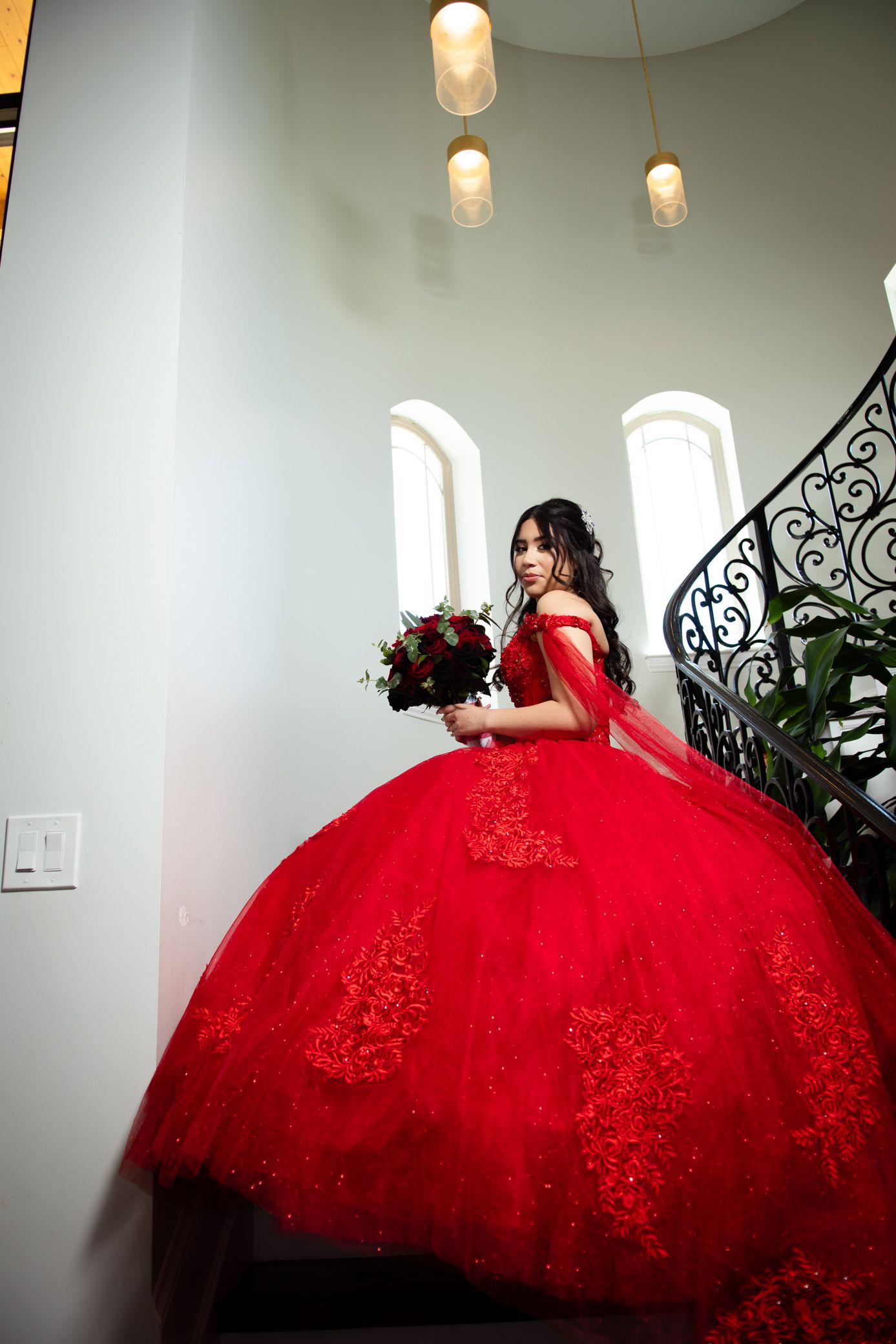 Young woman in red off-shoulder quinceañera ball gown descends grand staircase holding floral bouquet.