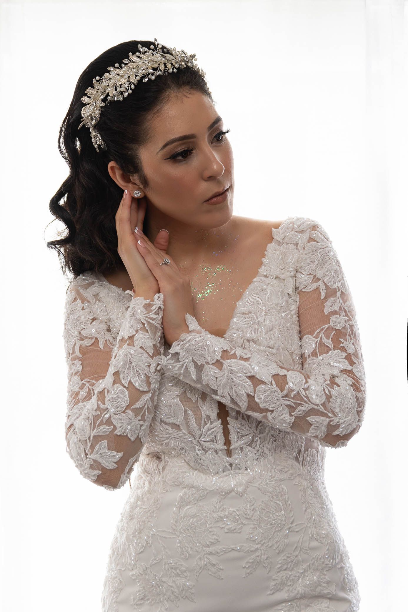 Bride in long-sleeve lace wedding dress adjusts earring while wearing a floral hair accessory.