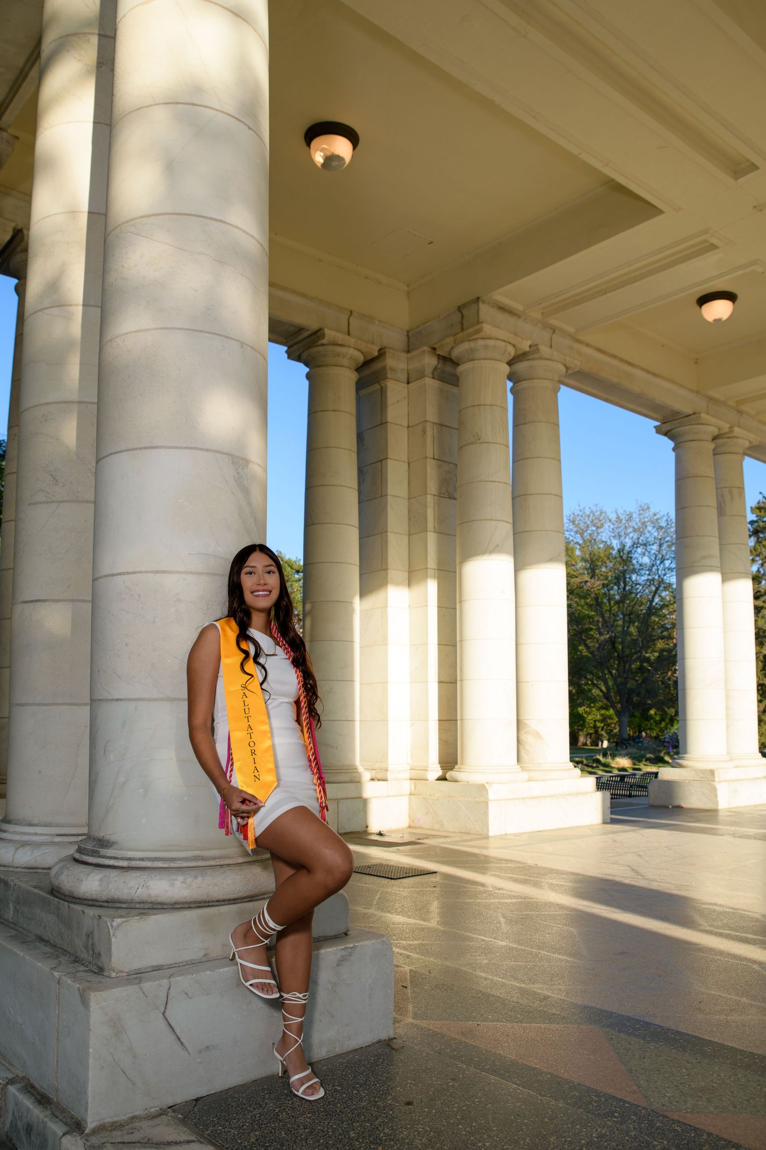 Young woman in white dress and graduation stoles leaning against marble columns in sunlight.