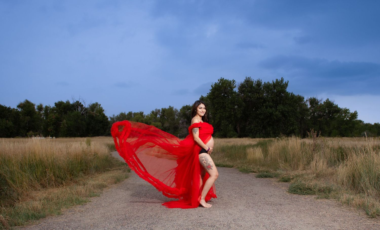 Pregnant woman in dramatic flowing red dress posing on a rural dirt path in open field.