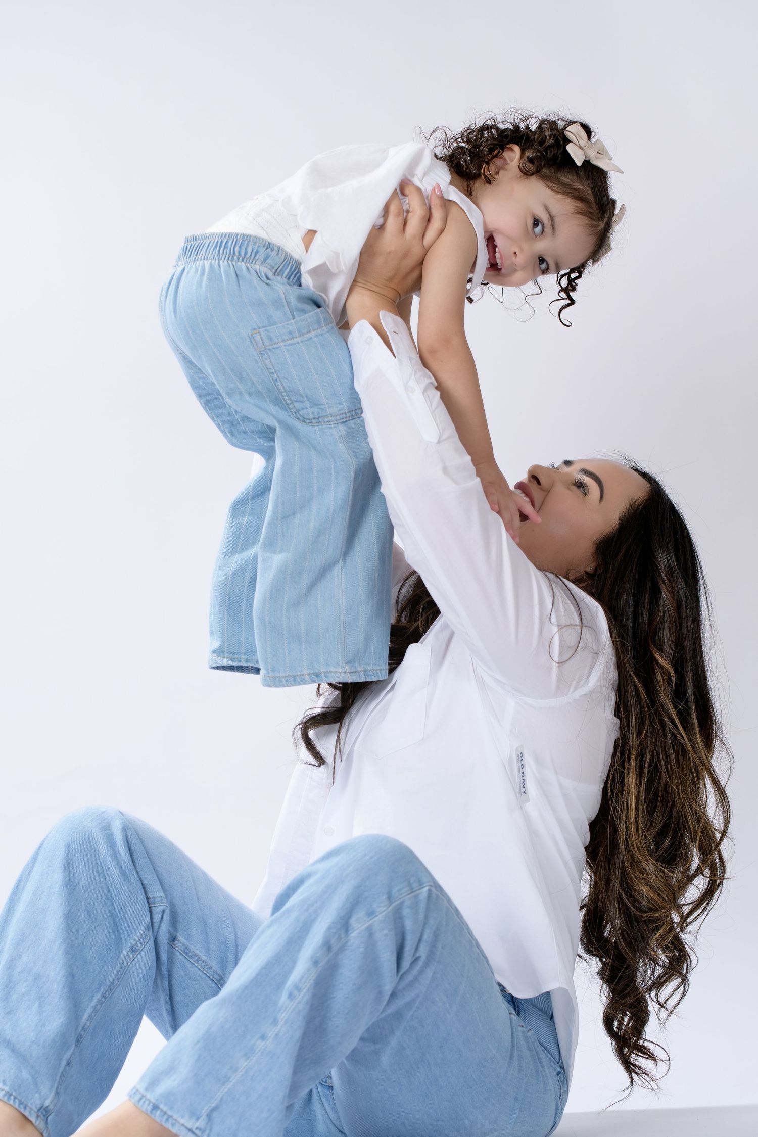 A mother lifts her curly-haired toddler girl playfully while both wear white tops and blue jeans.