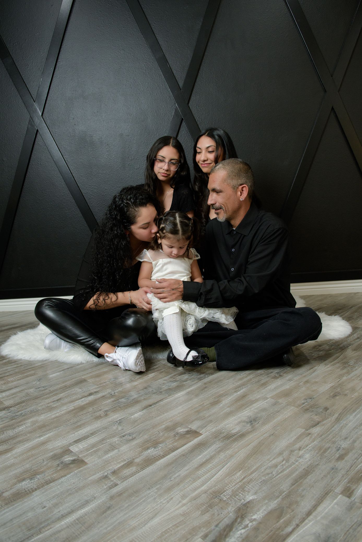 Family of five in black outfits sitting on fur rug against dark geometric wall, focusing on toddler in white dress.