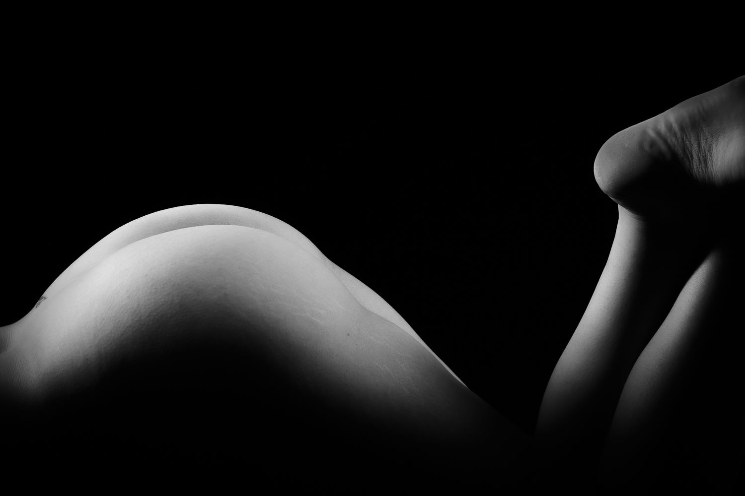 Minimalist black and white fine art bodyscape photograph highlighting feminine curves against dark background.