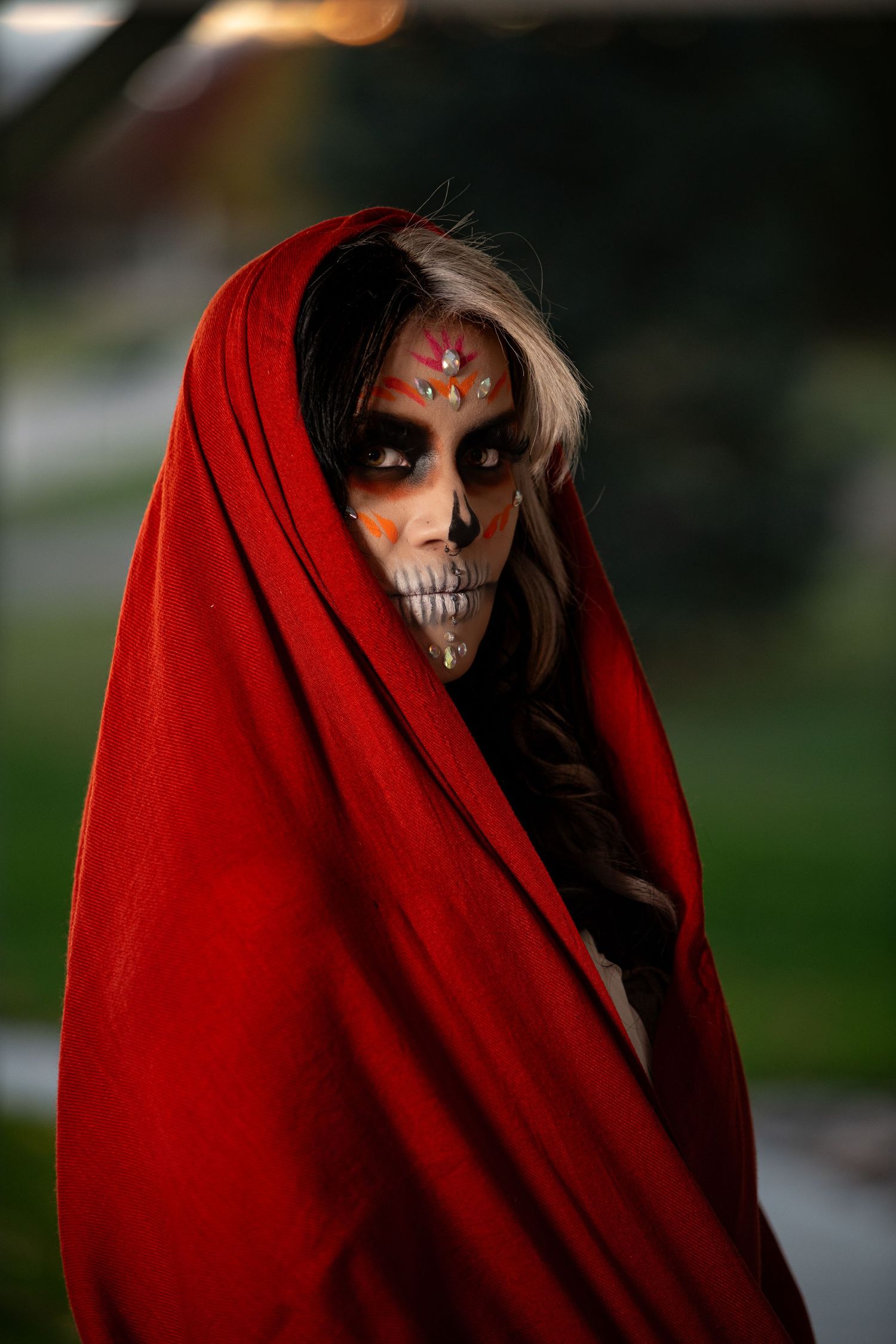 Woman draped in red cloth with detailed sugar skull makeup in moody outdoor portrait.