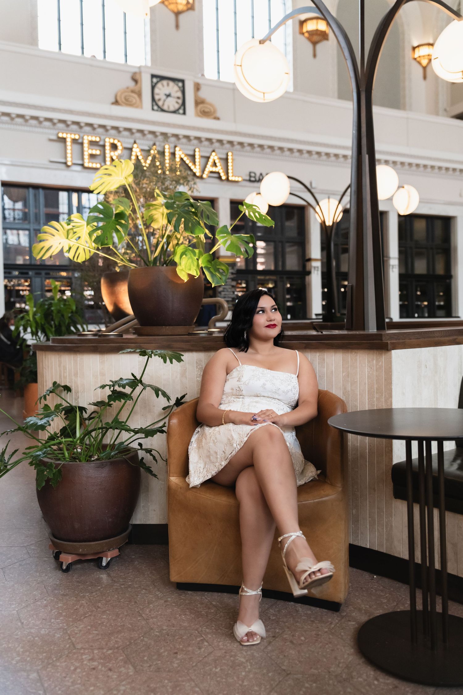 An elegant woman in a white mini dress sits in a leather chair inside a stylish terminal bar.