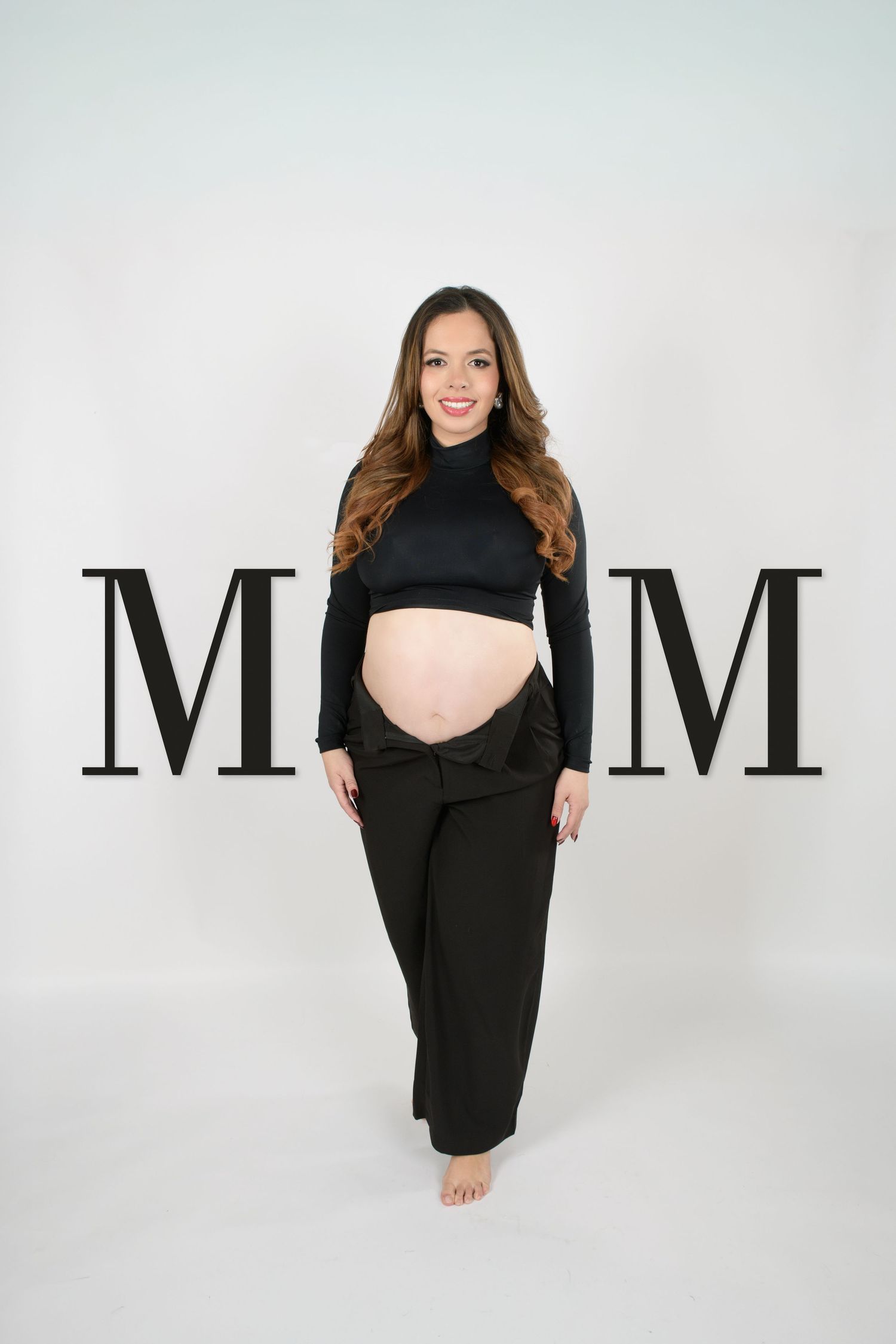 Smiling pregnant woman in black crop top and pants poses between large MOM letters in studio.