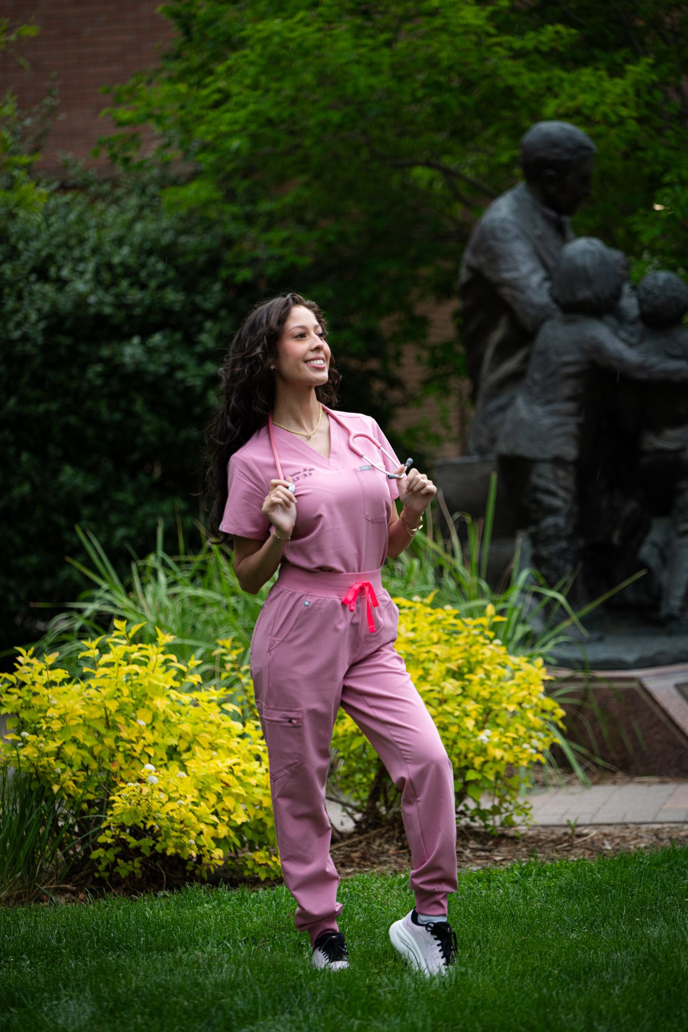 Happy female healthcare worker in pink scrubs with stethoscope posing in lush green garden.