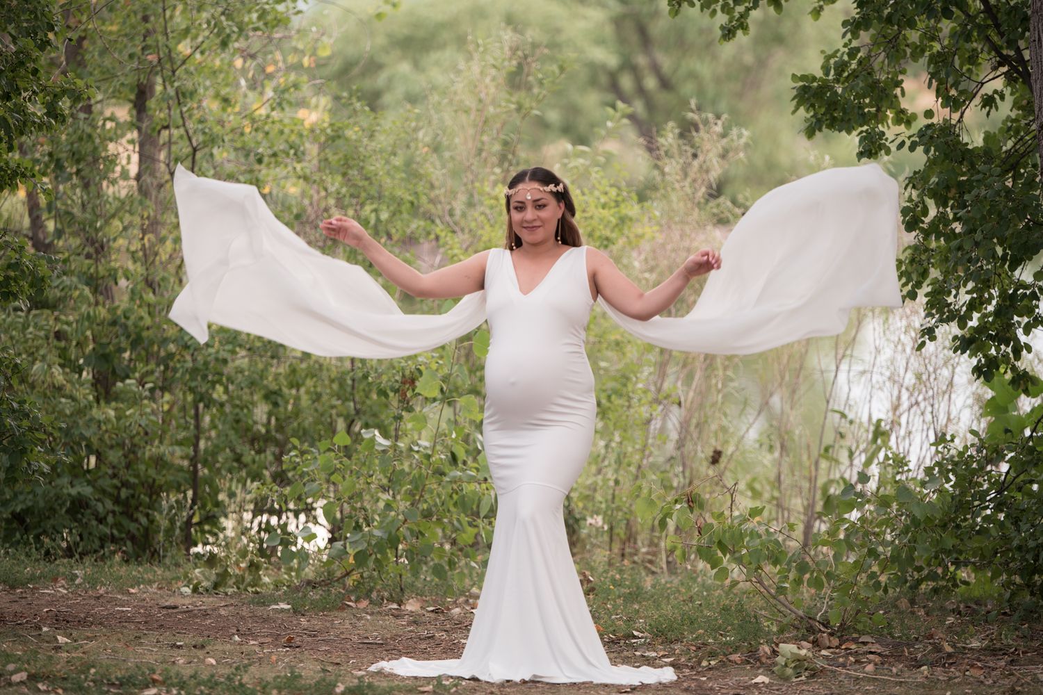 Pregnant woman in white fitted gown with flowing fabric arms outstretched in lush garden.