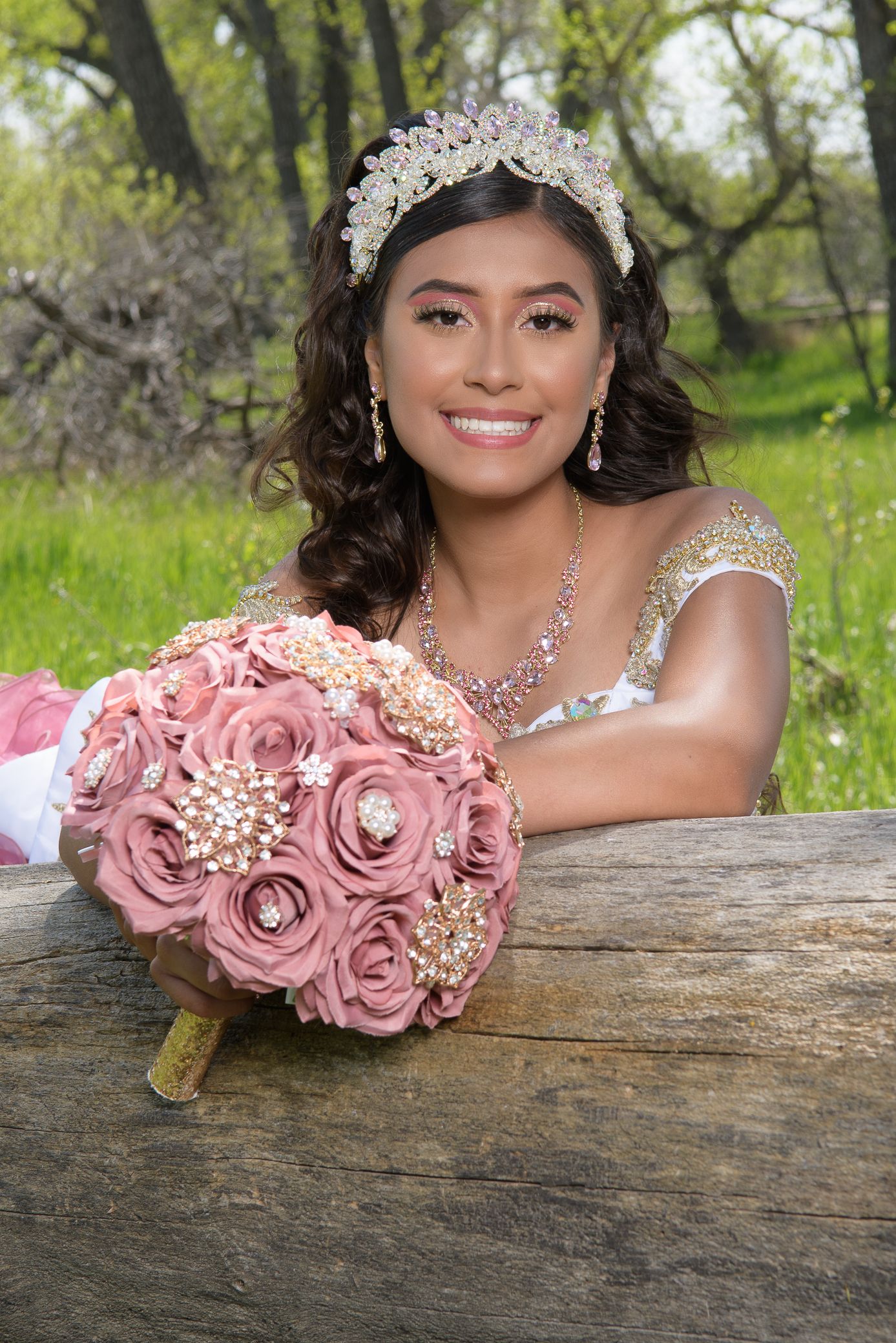 Smiling young woman in gold quinceañera dress holds pink rose brooch bouquet on rustic log.