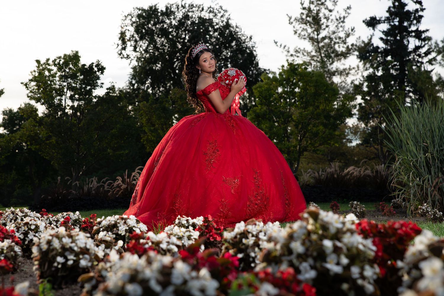 Young woman in red ball gown with tiara poses gracefully among colorful garden flower beds at dusk.