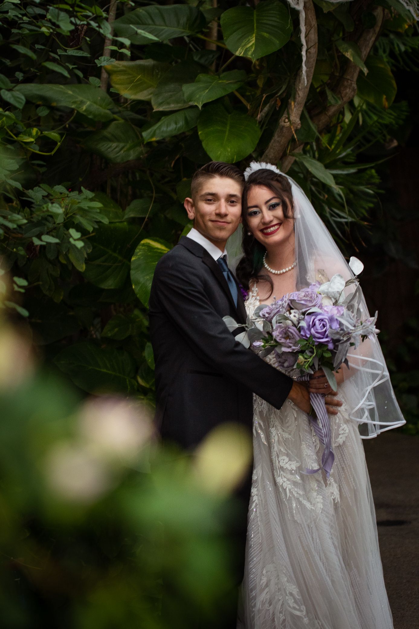 Smiling bride and groom pose together in a lush garden, bride holding a purple rose bouquet.